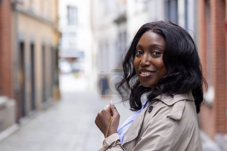 Cette image capture une jeune femme noire profitant d'un moment dans une ruelle européenne tranquille. Son expression joyeuse est complétée par sa tenue élégante, qui comprend un trench-coat beige et un bleu