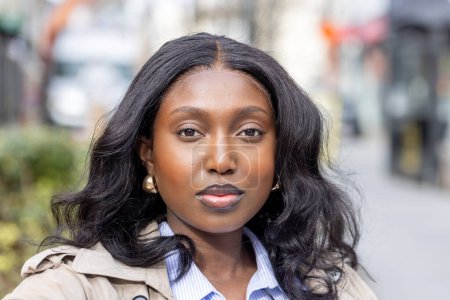 Photo for The photograph captures a young black woman with a confident and relaxed expression. She is dressed in a blue-striped button-down shirt and a beige trench coat, suggesting a professional yet - Royalty Free Image