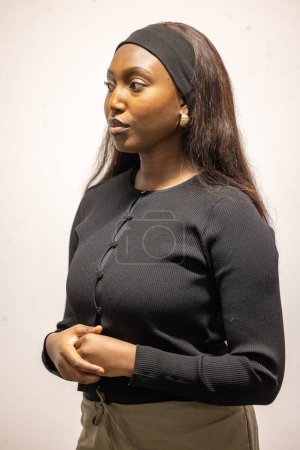 A young African woman stands against a light neutral background, exuding confidence and poise. She is dressed in a simple yet elegant black cardigan and her long hair is styled down, complemented by a