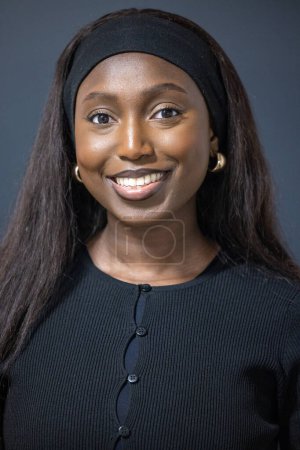 A close-up portrait of a radiant young African woman against a dark grey background. Her smile is engaging and joyful, highlighting her warm, friendly personality. She wears a stylish black headband