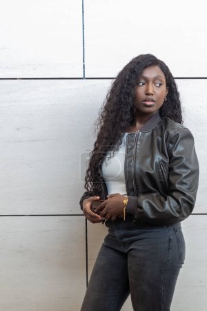 This image showcases a young African woman in urban casual attire, leaning confidently against a concrete wall. She dons a chic leather jacket over a simple white top, paired with classic denim jeans