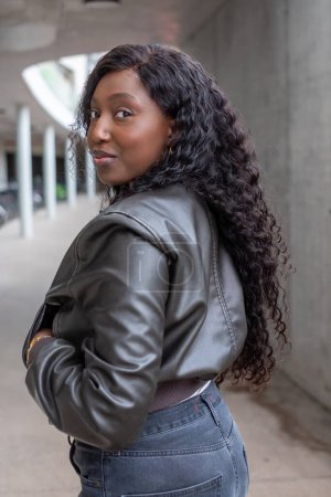In an urban setting, a confident African woman gives a playful over-the-shoulder look. Her curly hair flows freely, complementing the modern edge of her leather jacket. The soft focus on the concrete