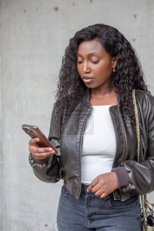 This image captures a young African woman focused intently on her smartphone. Standing against a textured concrete wall, she is styled in a modern leather jacket layered over a crisp white top, with