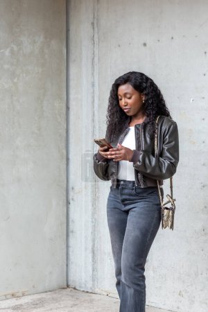 Captured in a moment of connectivity, a modern young woman stands engrossed in her smartphone against the minimalist backdrop of an urban concrete wall. Dressed in a fashionable leather jacket and