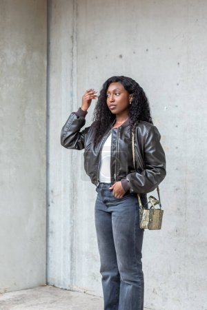 In this contemporary urban portrait, a stylish young woman leans casually against a concrete wall. Shes fashion-forward in a sleek leather jacket and grey jeans, her hair falling in natural waves