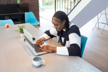In a modern and bright home setting, a woman is captured in a moment of joy as she works on her laptop. The natural light from the window illuminates her space, which is neatly organized with a coffee