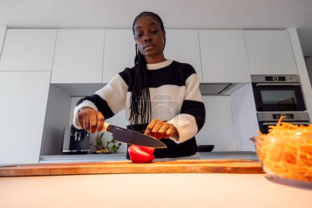 Une jeune femme est représentée dans un état de concentration profonde en train de couper un poivron rouge sur une planche à découper en bois. Les cuisines esthétiques contemporaines sont soulignées par la simplicité et