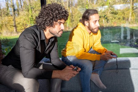 Dos amigos están profundamente enfocados mientras juegan videojuegos en una sala de estar llena de luz natural. La intensidad en sus rostros refleja la inmersión en el juego, con controladores de juego en la mano. El