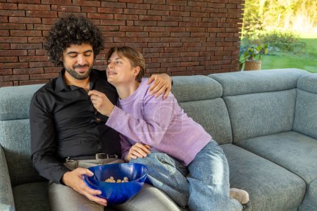 A couple enjoys a comfortable and intimate moment on the couch, sharing a bowl of snacks in a living room that exudes warmth. The woman playfully teases the man, who responds with a smile, indicating