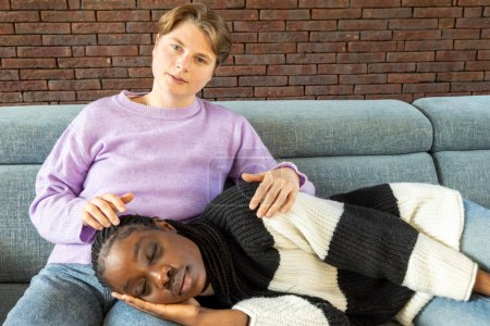 Photo for This tender scene depicts a lesbian gay couple, one woman resting with her head on the lap of her partner, who looks into the camera with a comforting, protective gaze. The casual setting of their - Royalty Free Image