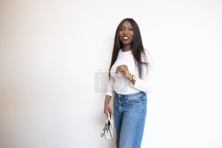 This vibrant image captures a smiling young Black woman dressed in a casual, stylish manner, standing against a plain white background. She wears a classic white top and blue jeans, accessorized with