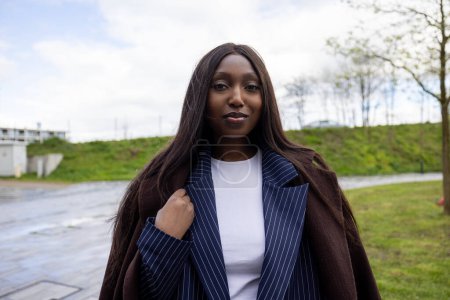The photograph depicts a self-assured young Black woman outdoors, wearing a stylish pinstripe blazer layered under a cozy brown coat. She stands with a gentle smile, subtly touching her coat, which