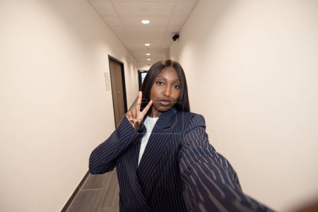 Capturing a spontaneous moment, this image features an animated young Black businesswoman taking a selfie in an office hallway. She makes a peace sign, adding a playful twist to her formal pinstripe