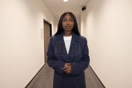 This photograph captures a poised young adult Black woman standing confidently in an office hallway. She sports a professional navy blue striped suit and white top, hands clasped in front of her. Her