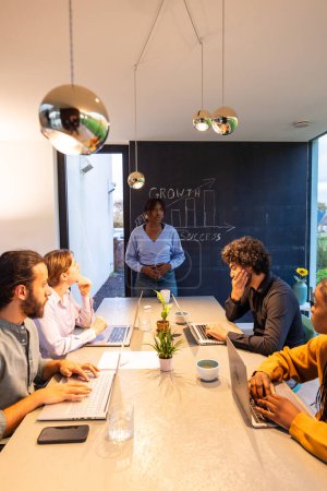 A diverse group of professionals gathers around a sleek white table in a contemporary office. The focus is on a confident African American woman standing by a blackboard with growth and success