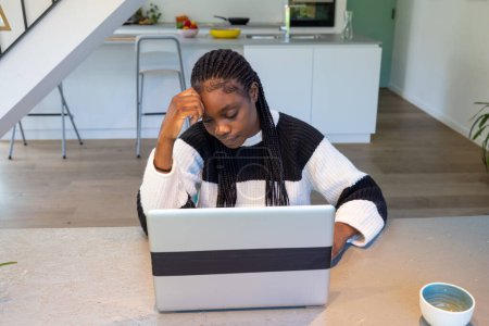 Une scène intime d'une jeune femme noire profondément concentrée sur son travail à la maison. Elle est assise à une table propre et moderne, absorbée dans son écran d'ordinateur portable avec une expression réfléchie, symbolisant le dévouement