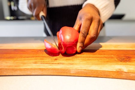 This image highlights the hands of a person skillfully slicing a ripe red bell pepper on a wooden cutting board. The action is captured mid-motion, emphasizing the freshness of the produce and the