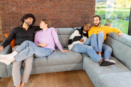 The image offers a glimpse into a relaxed moment among friends lounging on a plush sofa. Set in a room with a warm brick wall and a view to the garden, the scene exudes warmth and comfort. The group