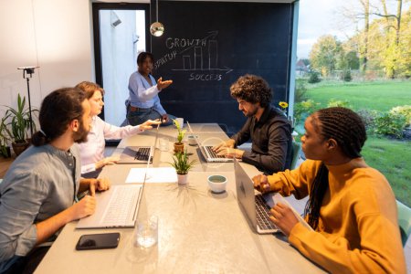 Dieses Foto fängt einen dynamischen Moment innerhalb eines vielfältigen Teams ein, das an einem Geschäftstreffen teilnimmt. Das Ambiente ist ein moderner, in natürliches Licht getauchter Büroraum mit freiem Blick ins Grüne. A