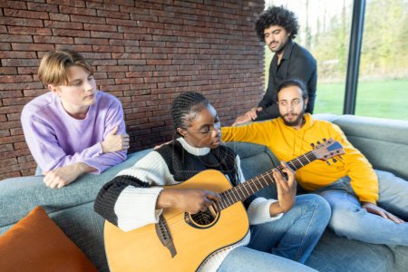 This image warmly depicts a moment of musical exchange among friends in a comfortable, homely setting. A woman is intently playing the guitar while her friends watch and listen, creating a casual and
