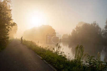 Dieses heitere Bild zeigt eine einsame Person, die einen nebelbedeckten Pfad am Flussufer hinuntergeht, wobei das sanfte Tageslicht einen Heiligenschein durch die Bäume erzeugt. Ein vom Nebel teilweise verdunkeltes Haus ist zu sehen