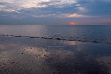 The image conveys a serene sunset at low tide, where the receding water leaves wet sand behind, mirroring the dwindling light. The sun, a fiery orb, dips towards the horizon, its warm glow contrasting