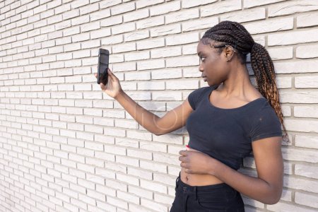 In this image, a young African woman is positioned in profile, holding up her smartphone to take a selfie. She is set against a backdrop of a white brick wall, creating a monochromatic background