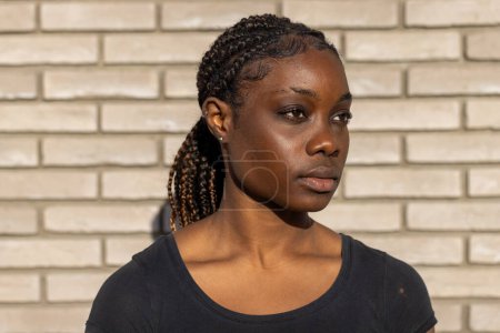 Eine junge Afroamerikanerin steht kontemplativ vor einer weißen Backsteinwand. Ihre Haare sind sauber zu Zöpfen gestylt, die sich über eine Schulter ziehen, und sie trägt ein einfaches schwarzes T-Shirt, das