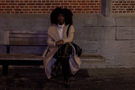 The photograph depicts a woman seated contemplatively on a bench, her presence engaging yet isolated against the urban backdrop of a brick wall. The nighttime setting envelopes her in a cloak of