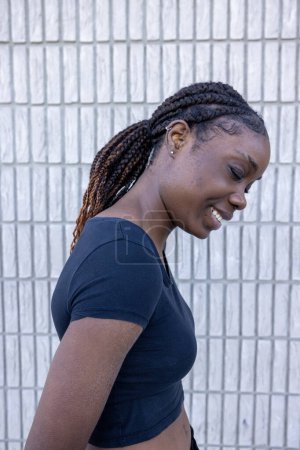 Captured in profile, this image depicts a young African woman with a joyful smile, looking downward. Her hair is styled in elegant braids that drape over her shoulder, and shes wearing a casual black