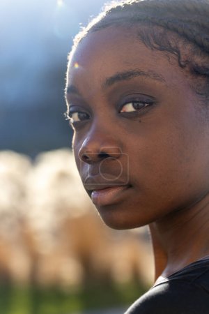 Este retrato de cerca presenta a una mujer africana serena con luz solar iluminando suavemente su rostro sobre un fondo natural borroso. El cálido resplandor crea un efecto halo alrededor de su silueta. Ella...