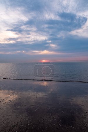 This photograph captures the tranquil beauty of early morning with the sunrise gently gracing the oceans horizon. The quiet play of light and shadow across the clouds and the soft reflection on the