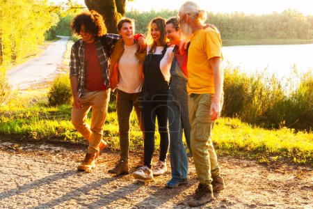 The image captures a candid moment among a diverse group of five friends as they enjoy a leisurely walk along a scenic lakeside path at sunset. The warm sunlight filters through the trees, casting a