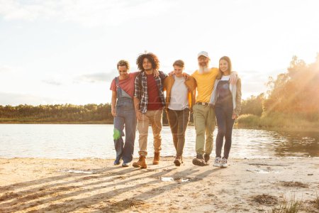 A bonded group of five strolls by the waters edge as the sun sets, casting a soft glow on their casual outing. Close-knit Friends Walking Together Along a Lakeshore at Dusk. High quality photo