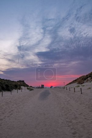 This image captures the allure of a beach at dusk, where a sandy path, flanked by dunes and grass, leads the eye to the sea. The sky above is a magnificent display of natures palette, with deep