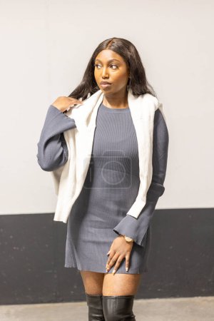 This image captures a young woman with a pensive expression, standing against a plain background that emphasizes her attire. She sports a snug grey dress paired with a relaxed-fit white blazer slung