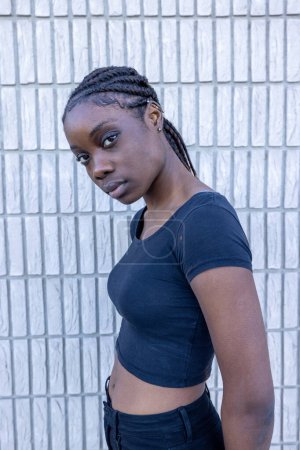 Una joven mujer africana está de pie en una pose casual pero elegante sobre un fondo de ladrillo blanco, su mirada girando ligeramente fuera de cámara, lo que sugiere introspección. El top negro apropiado y jeans