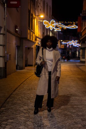 Una mujer toma las calles de la ciudad en una noche de invierno crujiente, su camino iluminado por las luces festivas encadenadas arriba. Su moda es una mezcla de calor estacional y estilo moderno, un abrigo largo en capas