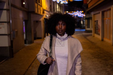 This evocative photograph showcases a stylish woman walking alone on a cobblestone street at dusk. The warm glow from the streetlights casts a soft light on her, complementing the festive string