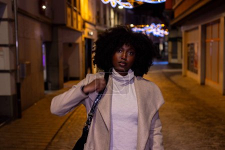 This compelling image captures a young woman confidently walking through a city street at night. The holiday lights softly illuminate the background, giving a festive mood to the urban setting. The