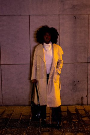 This image captures a striking pose of a woman against the minimalist setting of a concrete wall, under the nights ambient city light. Her attire, a chic coat over a turtleneck, paired with dark