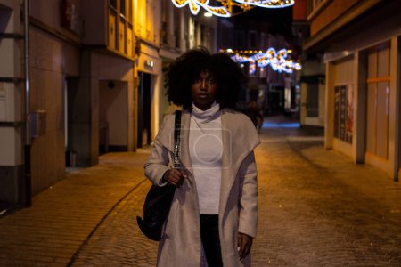 This striking image captures a confident woman walking towards the camera on an empty cobblestone street at night. Her bold afro and stylish outfit are illuminated by the soft glow of street lamps