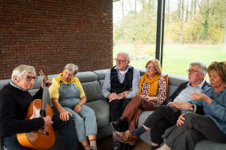 A heartwarming scene unfolds as a group of seniors gathers on a comfortable sofa in a well-lit room with large windows overlooking green scenery. One of them strums a guitar, bringing music to the mix