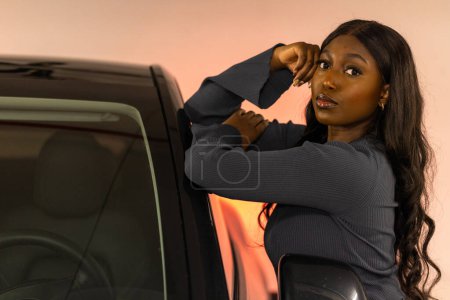 Eine balancierte Afroamerikanerin lehnt an ihrem Auto, ihre Hand liegt elegant auf dem Dach, als bereite sie sich auf eine Fahrt in den Sonnenuntergang vor. Die warmen Farbtöne, die die Szene umhüllen, deuten auf das Ende der