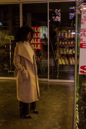 The warmth of a city evening is encapsulated in this image, where a woman stands by a shop window, the word SALE mirrored in bold letters. The festive lights twinkle in the background, suggesting a