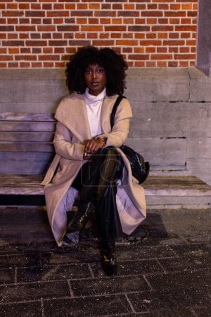 Dieses Bild fängt einen Moment der Ruhe ein und zeigt eine modebewusste Frau, die auf den Stufen der Stadt vor dem Hintergrund warmgetönter Ziegel sitzt. Ihre Pose ist entspannt und dennoch gelassen, verströmt einen Hauch von Lässigkeit
