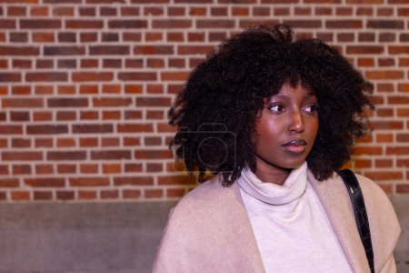 This image encapsulates a moment of introspection as a woman stands against the rich texture of a red brick wall. She appears to be caught in a moment of reverie, her profile to the camera, allowing