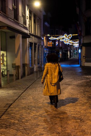 This image presents a solitary figure walking away along a cobblestone street under the soft glow of streetlights, with festive lights twinkling in the distance. The persons long coat and confident