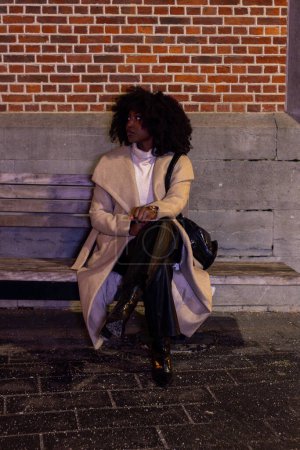 Auf diesem Bild sitzt eine modische junge Frau nachts allein auf den Stufen eines städtischen Gebäudes, ihre Silhouette wird von der städtischen Beleuchtung beleuchtet. Sie trägt ein stylisches Outfit, das die Weichheit