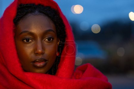 This evocative portrait features a young woman wrapped in a vibrant red scarf, her gaze penetrating and full of depth against the twilight backdrop. The scarf frames her face, drawing attention to her